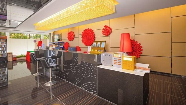 Un área de recepción moderna con un mostrador negro, dos taburetes de bar, una lámpara colgante amarilla y decoraciones florales rojas en la pared.