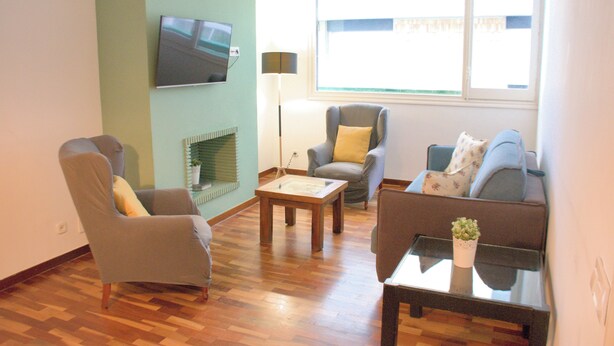 Una sala de estar con una televisión de pantalla plana montada en la pared, un sofá, una silla, una mesa de centro y una ventana con persianas.