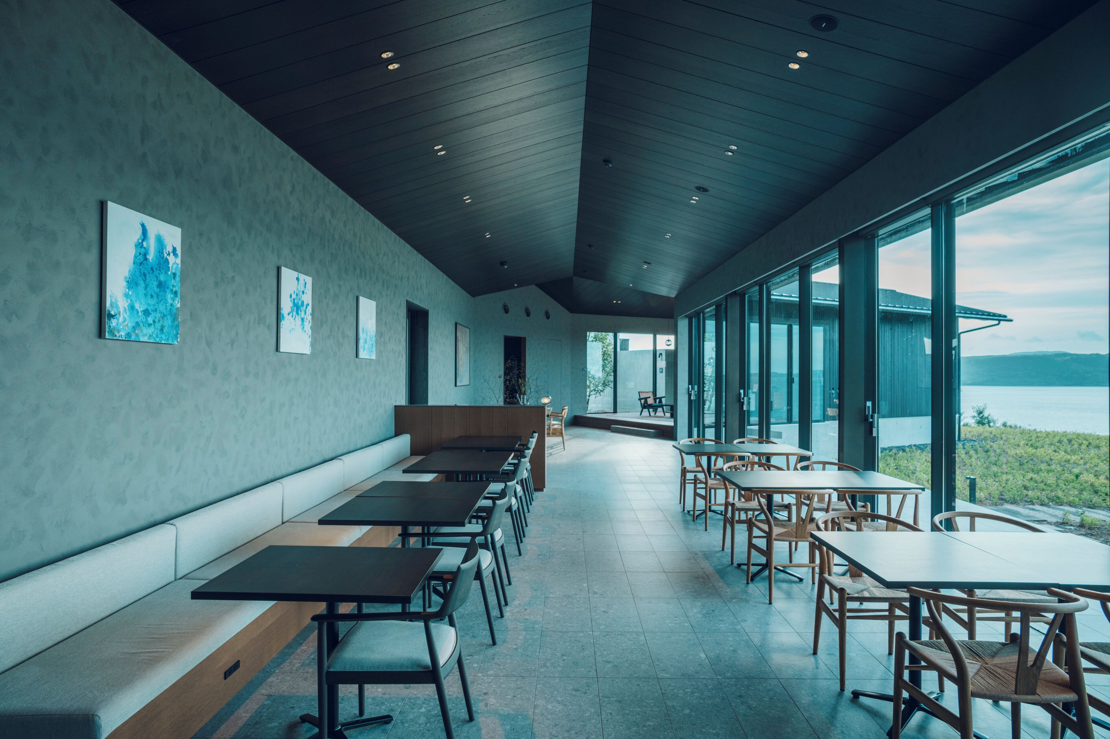 A modern dining area with long benches and tables, large windows offering a view of the outdoors.