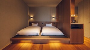 A modern hotel room with a wooden floor, two single beds with white bedding, a built-in wooden cabinet, and wall-mounted lighting. - Kikka Hirado (Hirado)