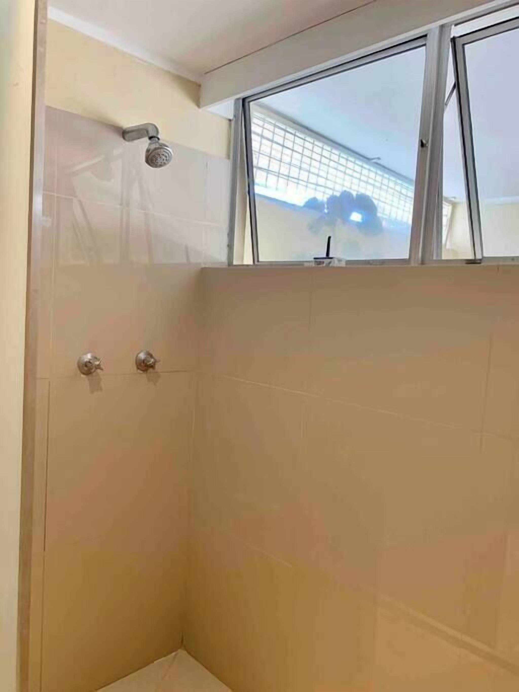 Un petit espace douche fermé, doté d’une fenêtre, d’un pommeau de douche et d’une tuyauterie visible.