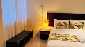 Habitación de hotel con cama, mesita de noche con lámpara, banqueta y ventana con cortinas.