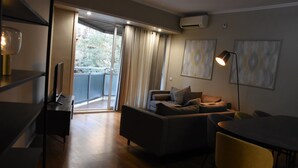 Ett modernt vardagsrum med en soffa, ett matbord och en gul stol. Det finns en balkong med gardiner och en luftkonditioneringsenhet på väggen.