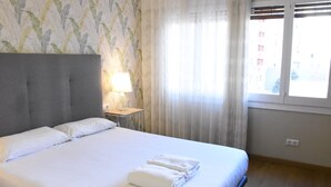 Ett hotellrum med en säng, ett nattduksbord med en lampa, ett fönster med gardiner och utsikt över byggnaderna utanför.
