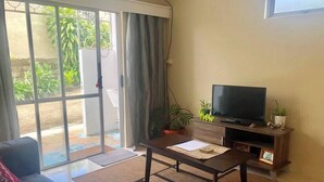 A living room with a sofa, coffee table, TV, and a sliding glass door leading to a balcony with plants.