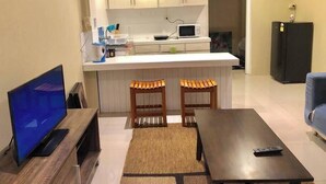 A compact living space with a TV, a coffee table, two wooden stools, a kitchen area with a microwave, and a refrigerator.