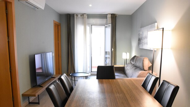 Una camera d'hotel moderna con un tavolo da pranzo in legno, sedie in pelle nera, una TV a schermo piatto, un divano e una lampada da terra.
