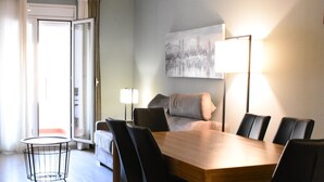 Una camera d'albergo moderna con zona pranzo, che include un tavolo e sedie in legno, un divano, una lampada e un quadro appeso al muro.