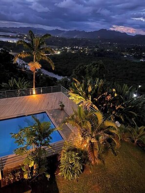 Zona de piscina amb un pont de fusta, envoltada de plantes tropicals i amb vista a un paisatge muntanyós al capvespre.