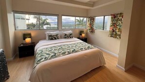Ein Schlafzimmer mit einem großen Bett, zwei Nachttischen, einem Beistelltisch und einem Fenster mit Blick auf Palmen und Berge.