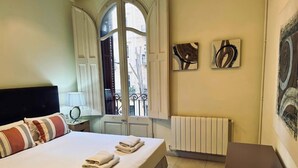 Ein Hotelzimmer mit Bett, Nachttisch, Fenster mit Vorhängen, Heizkörper und zwei Wandbildern.