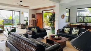 A spacious living area with a large sofa, a coffee table, and a ceiling fan. There are large windows providing natural light and a view of the outdoors.