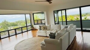 A spacious living room with a white sofa, a ceiling fan, and large windows offering a view of greenery.