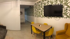 A modern living room with a grey sofa, a wooden dining table with yellow chairs, a flat-screen TV mounted on a leaf-patterned wall, and a painting on the wall. - TWO-BEDROOM APARTMENT (Barcelona)