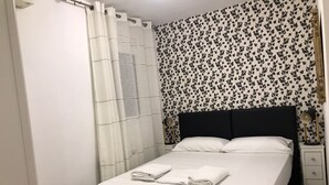 A neatly made bed with white linens and two pillows, a bedside table with a lamp, and a wall with a leafy pattern. - TWO-BEDROOM APARTMENT (Barcelona)