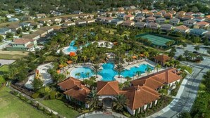 An aerial view of a residential area with a central swimming pool surrounded by houses, palm trees, and a tennis court.