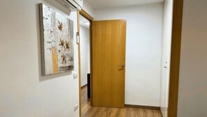 A hotel room with a wooden door, a white wall, and a wooden floor. There is a bed with a white blanket and a folded white towel on it.