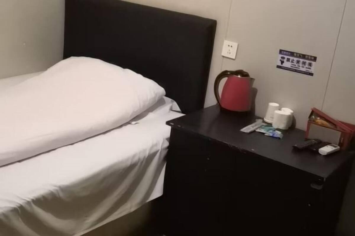 Een enkel bed met witte lakens, een zwart hoofdbord, een klein tafeltje met een rode waterkoker, enkele toiletartikelen en een bord aan de muur.