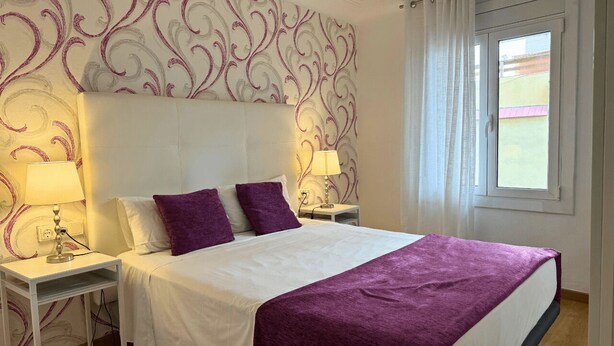 A neatly made bed with a white and purple bedspread, two purple pillows, and two bedside lamps on white nightstands. - 2 BEDROOM DUPLEX (Barcelona)