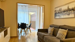 A living room with a sofa, a television, a dining table with chairs, and a sliding glass door leading to an outdoor area. - 2 BEDROOM DUPLEX (Barcelona)