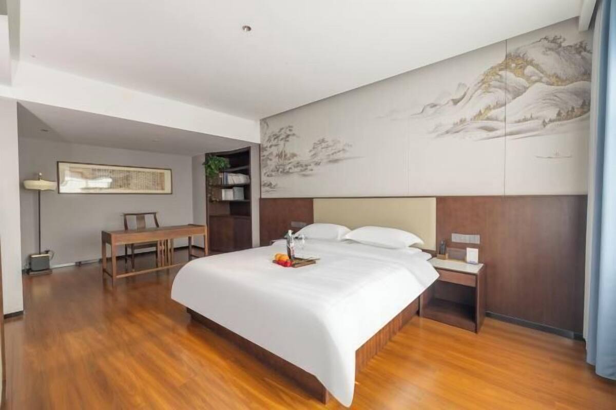 Una habitación de hotel con piso de madera, una cama con ropa blanca, una mesita de noche, un escritorio y una pared con un cuadro.