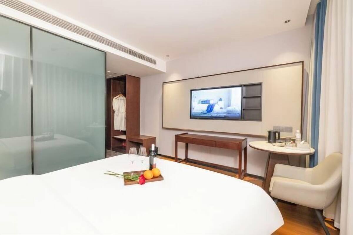 Una habitación de hotel con una cama grande, un televisor montado en la pared, una mesita con una botella y vasos, y una silla.