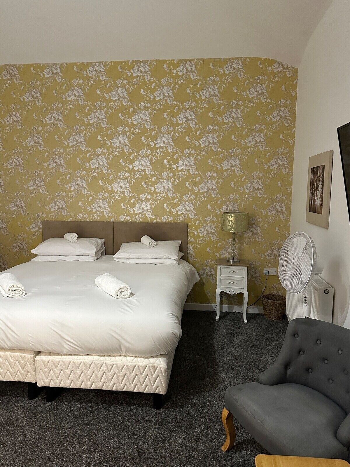 Habitación de hotel con una cama grande, una mesita de noche con lámpara, una silla y un ventilador de pared.