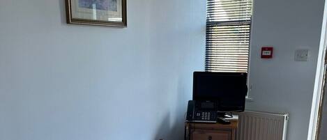 A neatly made bed with a rolled towel, a framed picture on the wall, a desk with a computer, and a window with blinds.