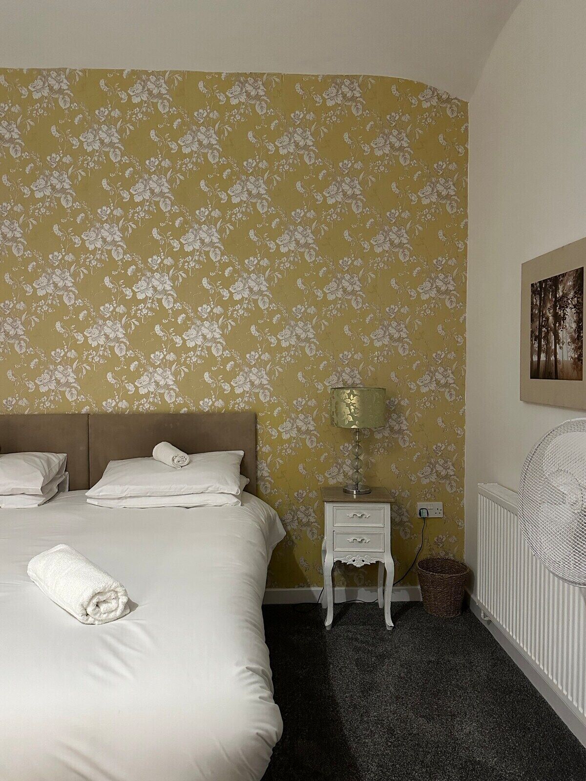 Ein Hotelzimmer mit blumigem Wandpapier, einem Bett mit weißer Bettwäsche, einer Nachttischlampe, einem weißen Heizkörper und einem gerahmten Bild an der Wand.
