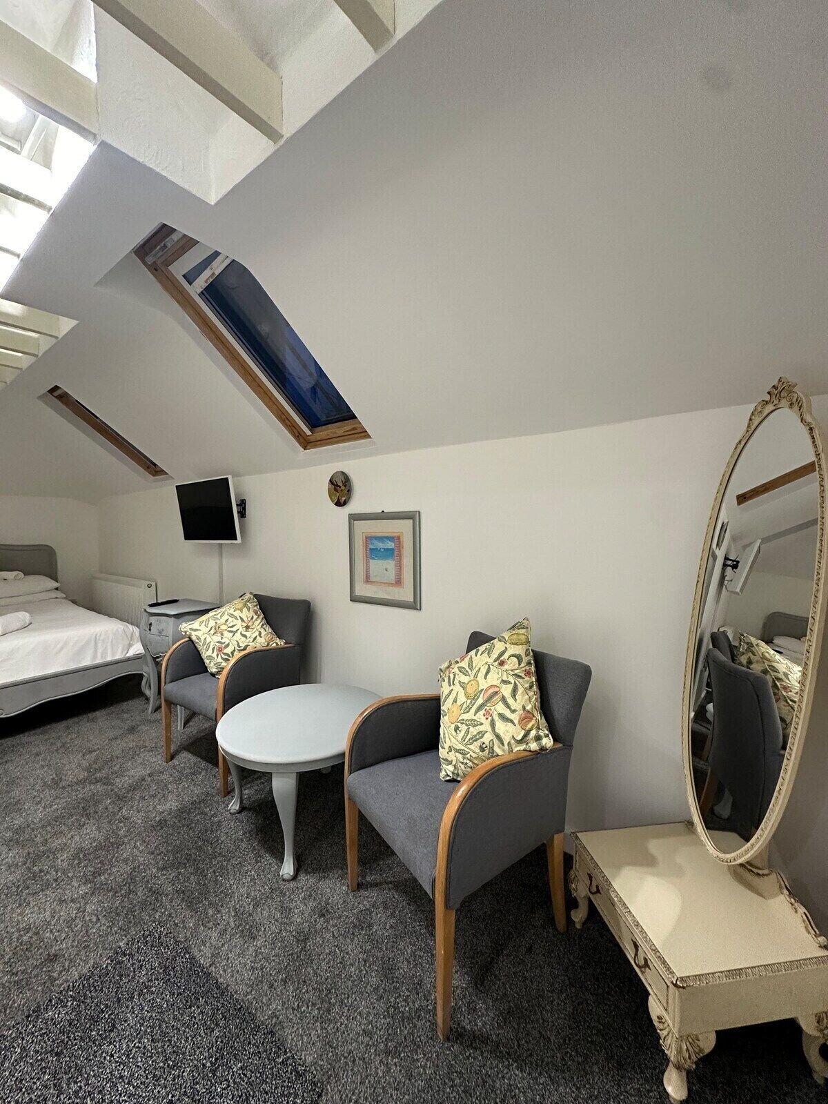 Une chambre d’hôtel compacte comprenant un lit, deux fauteuils, une petite table, un miroir rond et une lucarne au plafond.