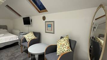 Une chambre d’hôtel compacte comprenant un lit, deux fauteuils, une petite table, un miroir rond et une lucarne au plafond.