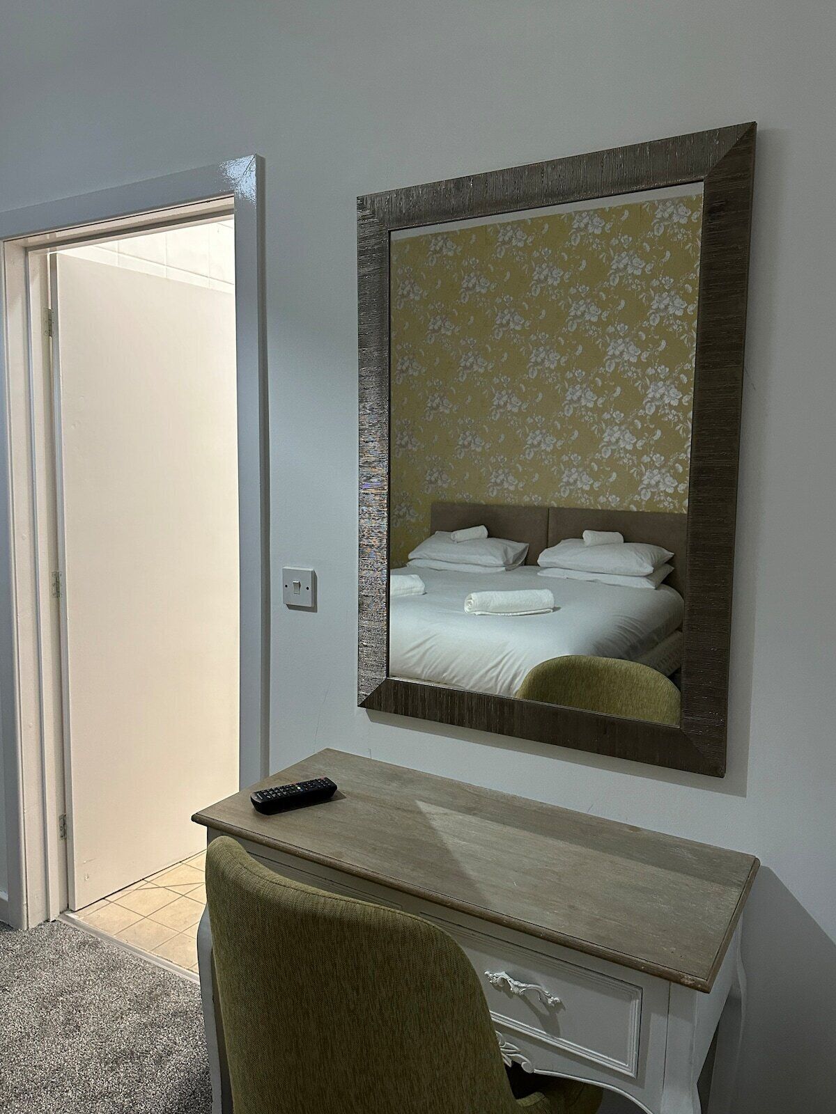 A hotel room with a bed, a desk with a chair, a mirror, and a door.