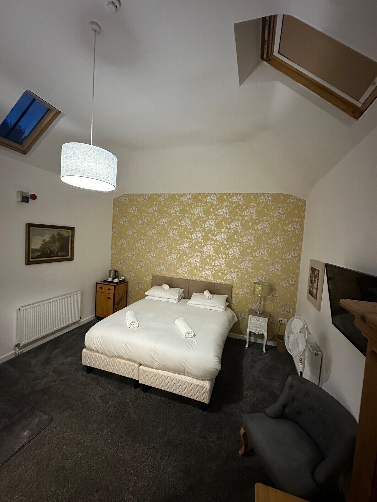 A bedroom with a bed, a bedside table, a lamp, a fan, and a picture on the wall.