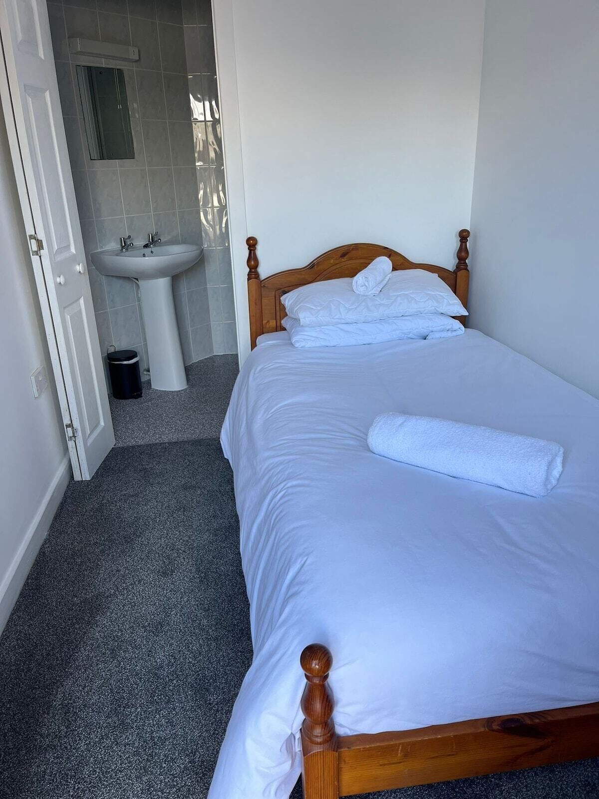 A neatly made bed with white linens and a wooden headboard, a white sink, and a small trash bin in a room with grey carpet.