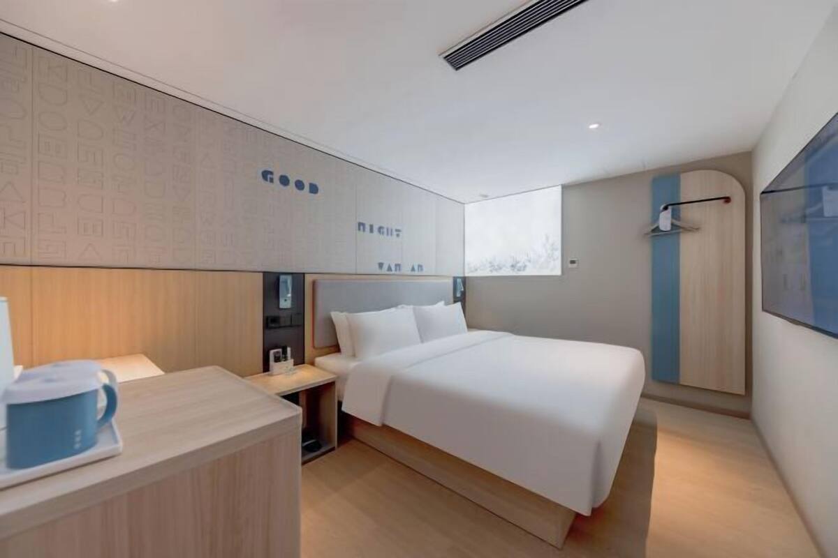 A modern hotel room with a large bed, a bedside table with a coffee maker, a wardrobe, and a large window.