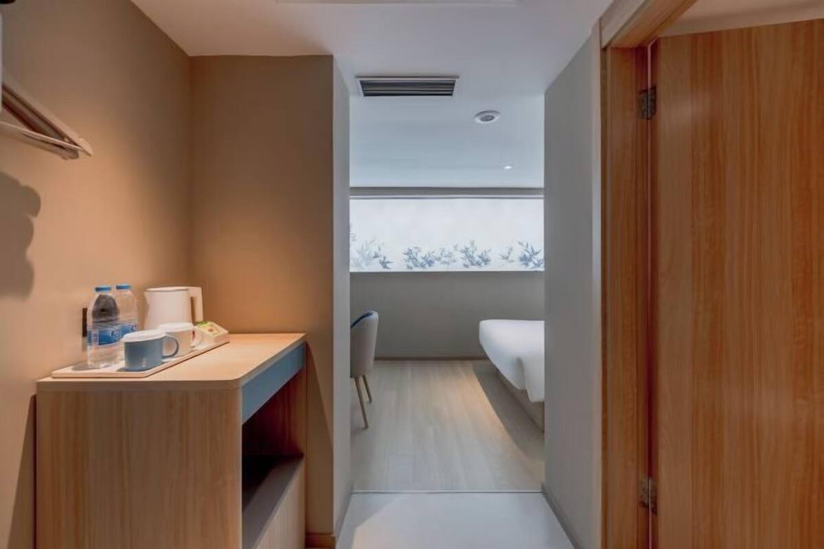 A compact room with a bed, a small desk with a cup and water bottle, a chair, and a window with a view of trees.