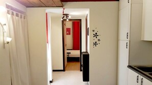 Um corredor estreito leva a um quarto com uma cama e uma cortina vermelha. Há uma parede branca com um desenho preto decorativo, um ventilador de teto e uma área de cozinha com armários e uma pia.