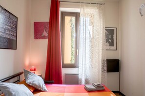 Une chambre à coucher avec un lit, des oreillers, une table de chevet avec une lampe, une fenêtre avec des rideaux et un tableau accroché au mur.
