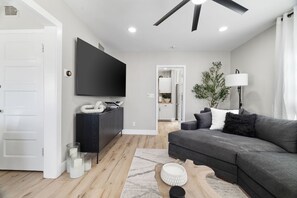 Una sala de estar moderna con un sofá gris, un televisor de pantalla plana, una mesa de centro de madera y una puerta blanca.