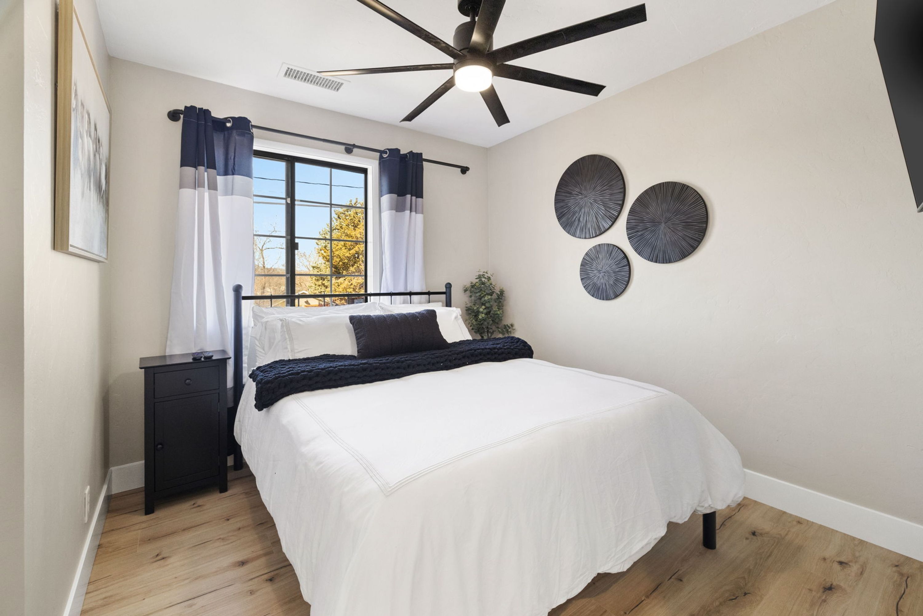 Une chambre à coucher comprenant un lit, une table de chevet, un ventilateur de plafond, une fenêtre avec des rideaux et des éléments de décoration murale.