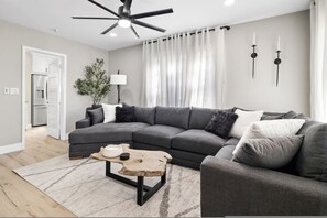 Una sala de estar moderna con un sofá modular, una mesa de centro de madera, un ventilador de techo y un ventanal con cortinas.