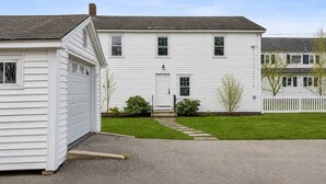 A two-story white house with a garage, a front door, and a small front yard with a pathway. - The Coastal Nest Stay for 5! (Scarborough)