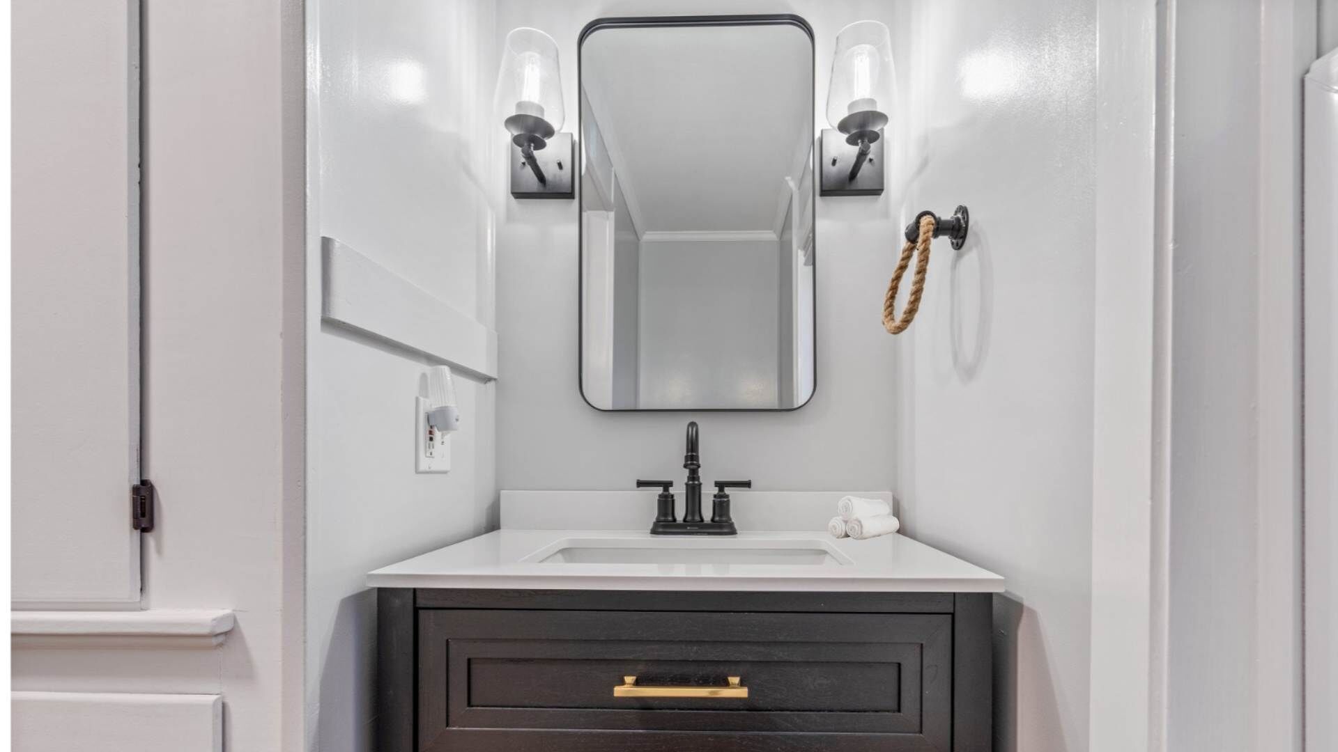 A bathroom with a white vanity, a dark cabinet below, a mirror above the sink, and two wall-mounted light fixtures.