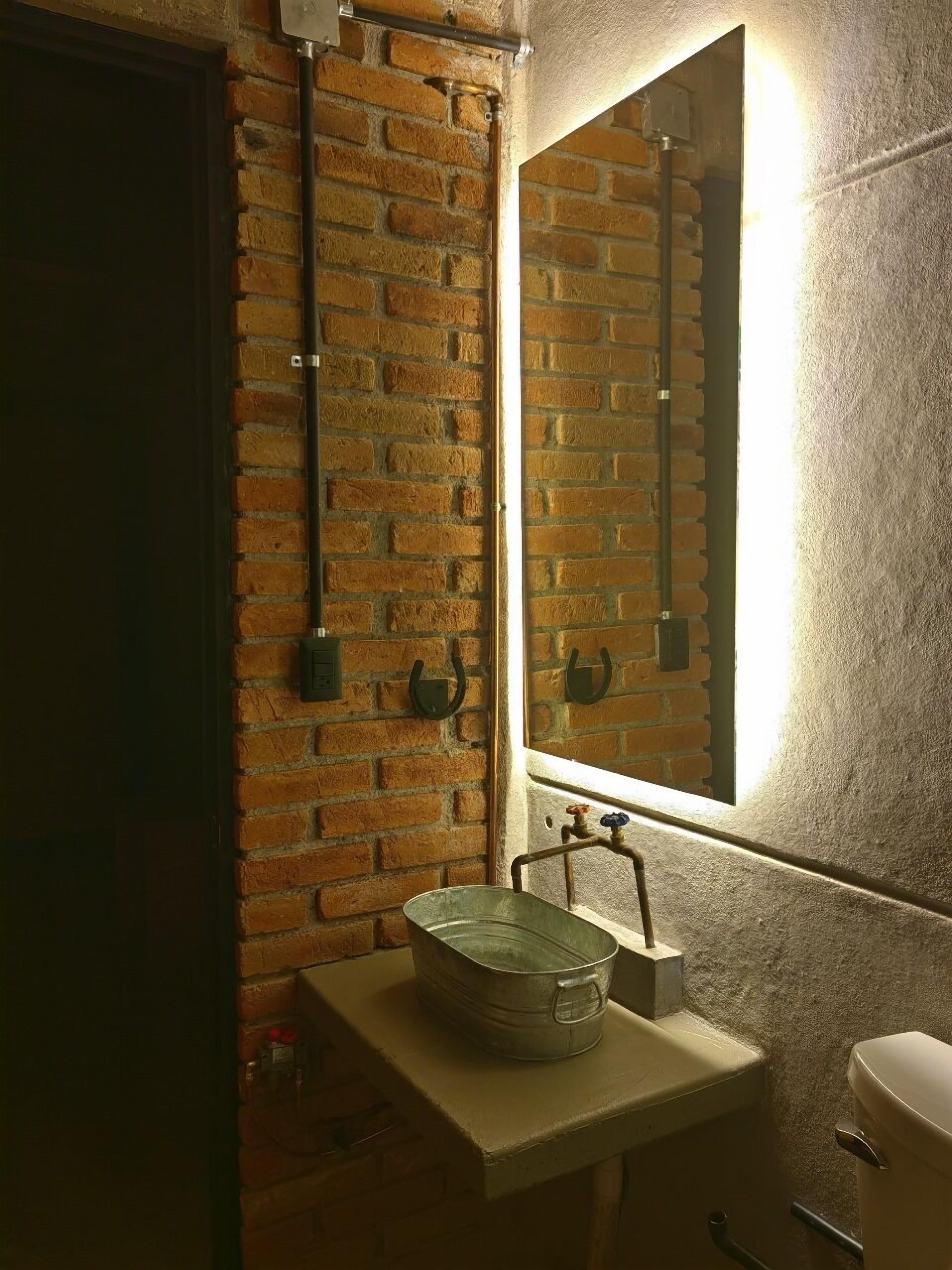 Et badeværelse med en murstensvæg, en rund metalvask, et spejl og et toilet.