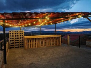 An outdoor patio with a wooden bar and wine storage, under a pergola with string lights, overlooking a cityscape at dusk. - Cabañas El Campo (Amealco)
