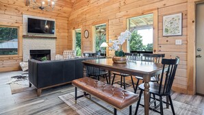 Un intérieur en bois avec une cheminée, une grande télévision, une table à manger avec des chaises et un banc.