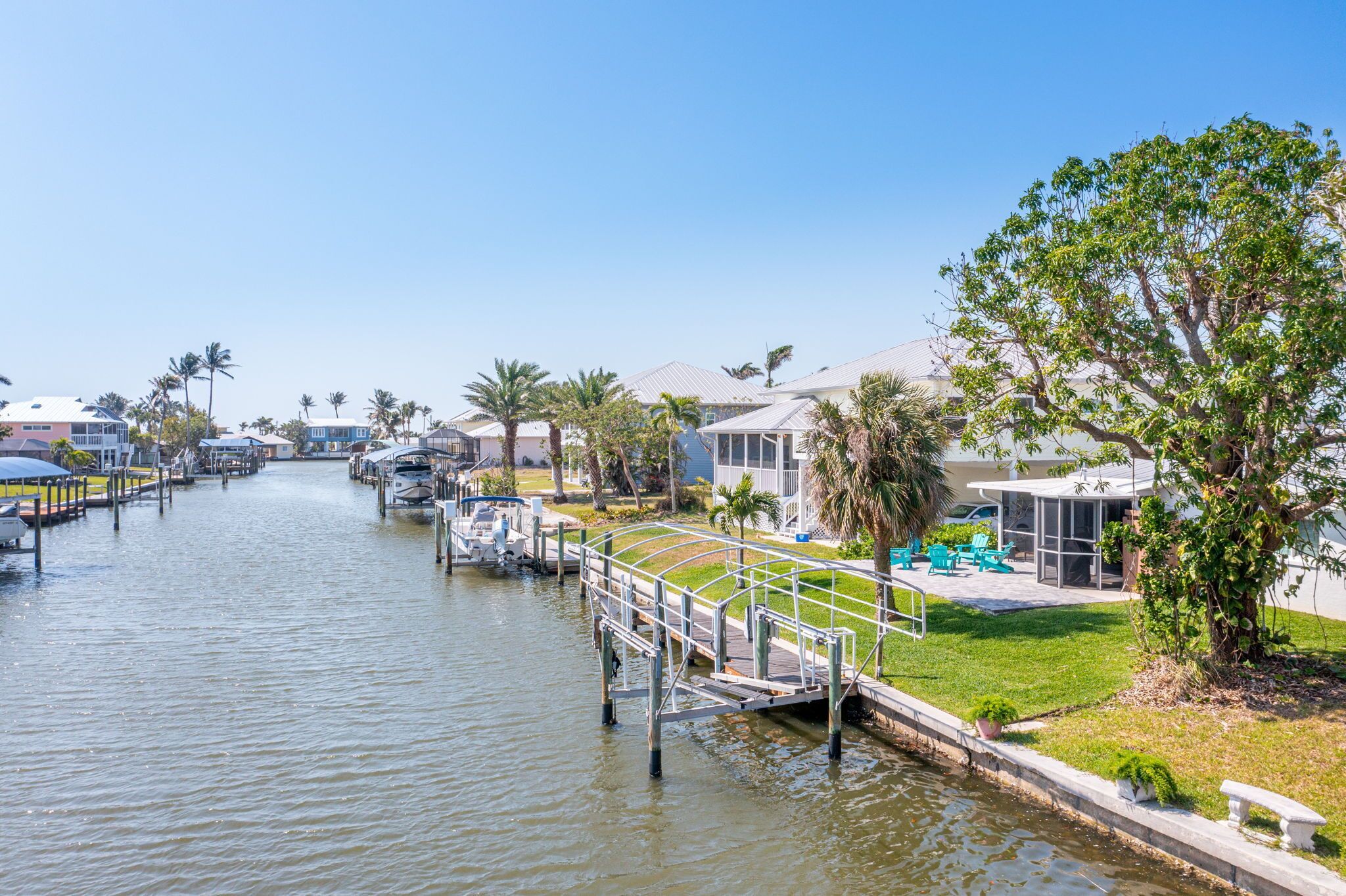A canal with boats docked, a pedestrian bridge, and houses with pools and greenery.