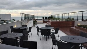 A rooftop terrace with round tables and chairs, a wooden bench area, and a view of the sea. - Apto 00203 Legacy Suites  (João Pessoa)