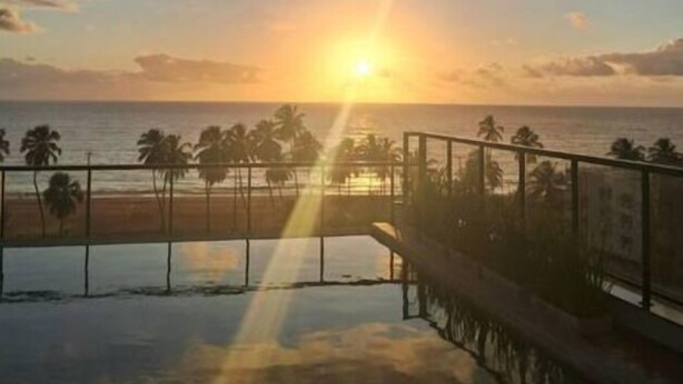 A sunset view from a balcony overlooking a beach with palm trees and a pool. - Apto 00203 Legacy Suites  (João Pessoa)