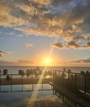 A sunset view from a balcony overlooking a beach with palm trees and a pool. - Apto 00203 Legacy Suites  (João Pessoa)